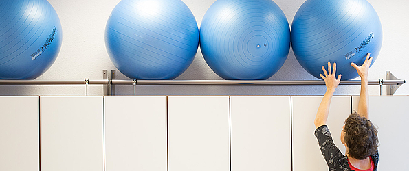 Eine Frau räumt auf und legt einen großen Gymnastikball zu weiteren Bällen auf ein Regal.
