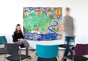 Eine Patientin und ein Patient sind in einem Zimmer mit mehreren Stühlen in einem Kreis, an der Wand hängt ein großes, gemaltes Bild.
